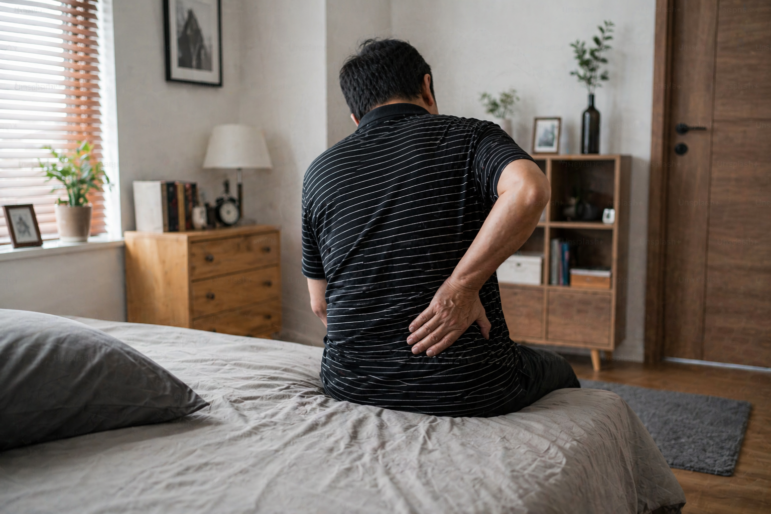 pria duduk di pinggir kasur sambil memegang pinggang